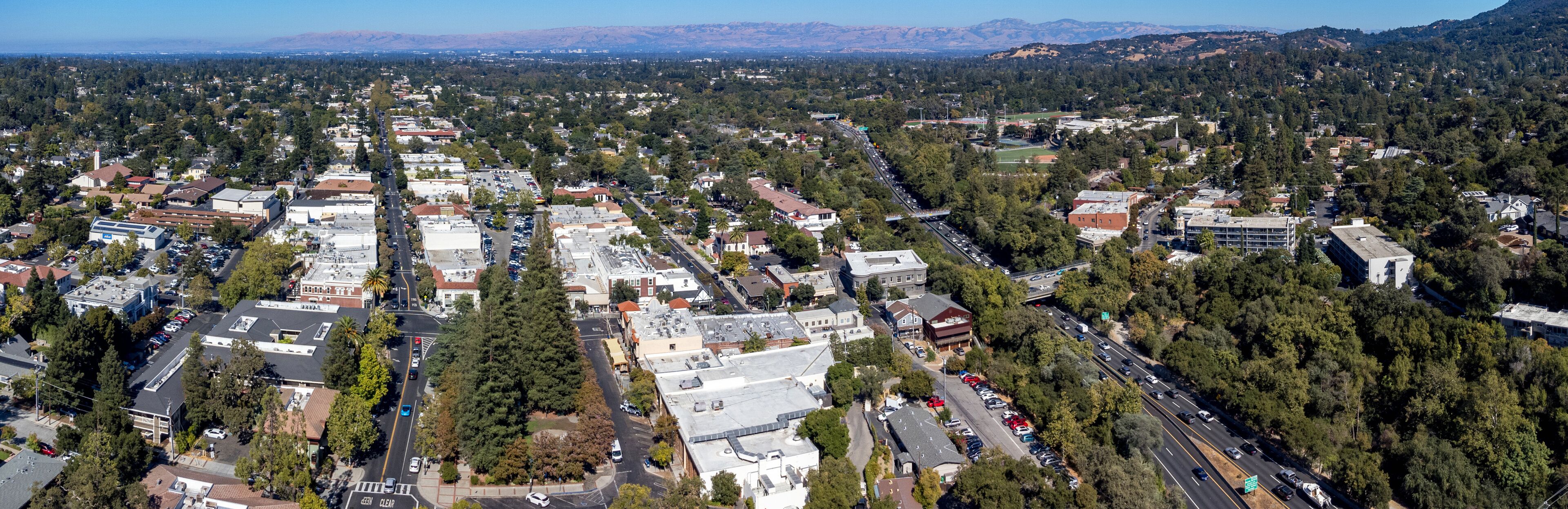 Aerial panoramic view of downtown Los Gatos with a mix of residential and commercial buildings. Highway 17 cuts through the suburban area with mountains in the background.