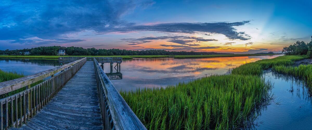 A wooden pier extending over calm water, surrounded by lush greenery and a sunset sky.