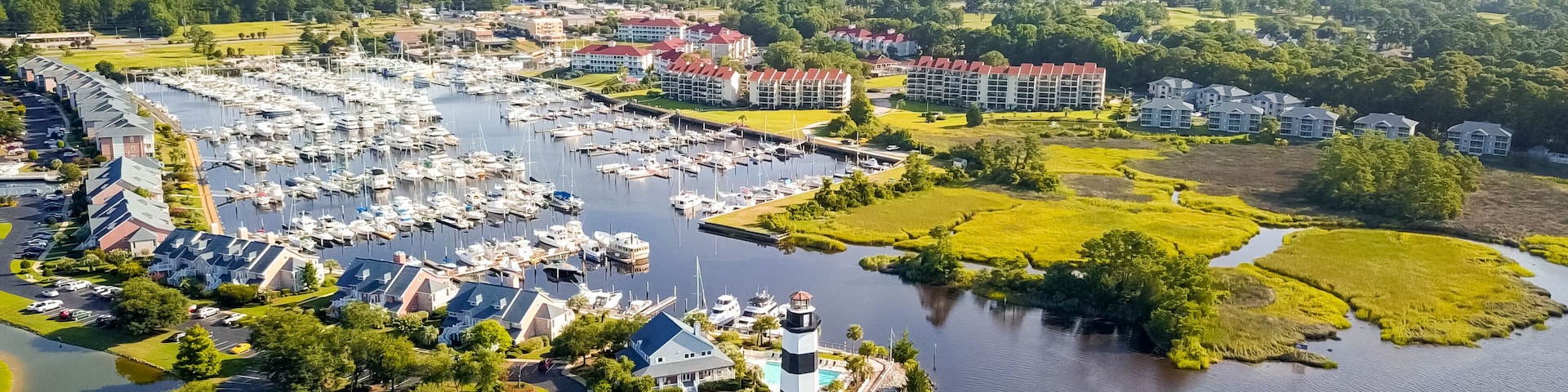 Vista aérea de uma marina com barcos, um farol e edifícios residenciais ao redor.