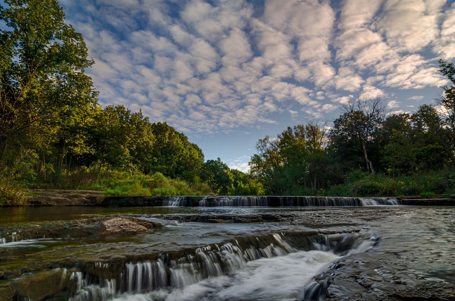 A waterfall cascading over rocks in a forested area with a cloudy sky.