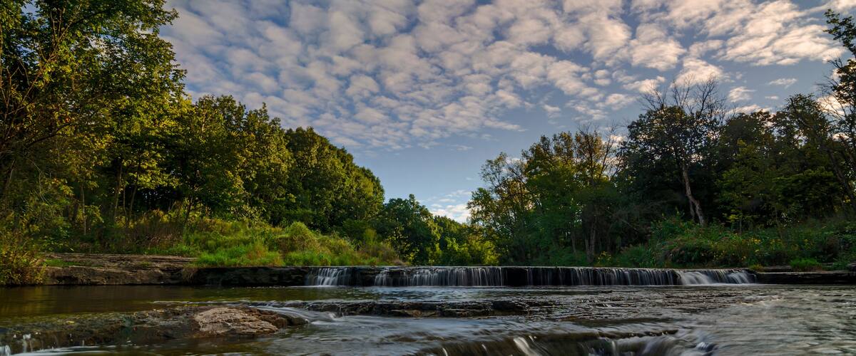 A waterfall cascading over rocks in a forested area with a cloudy sky.