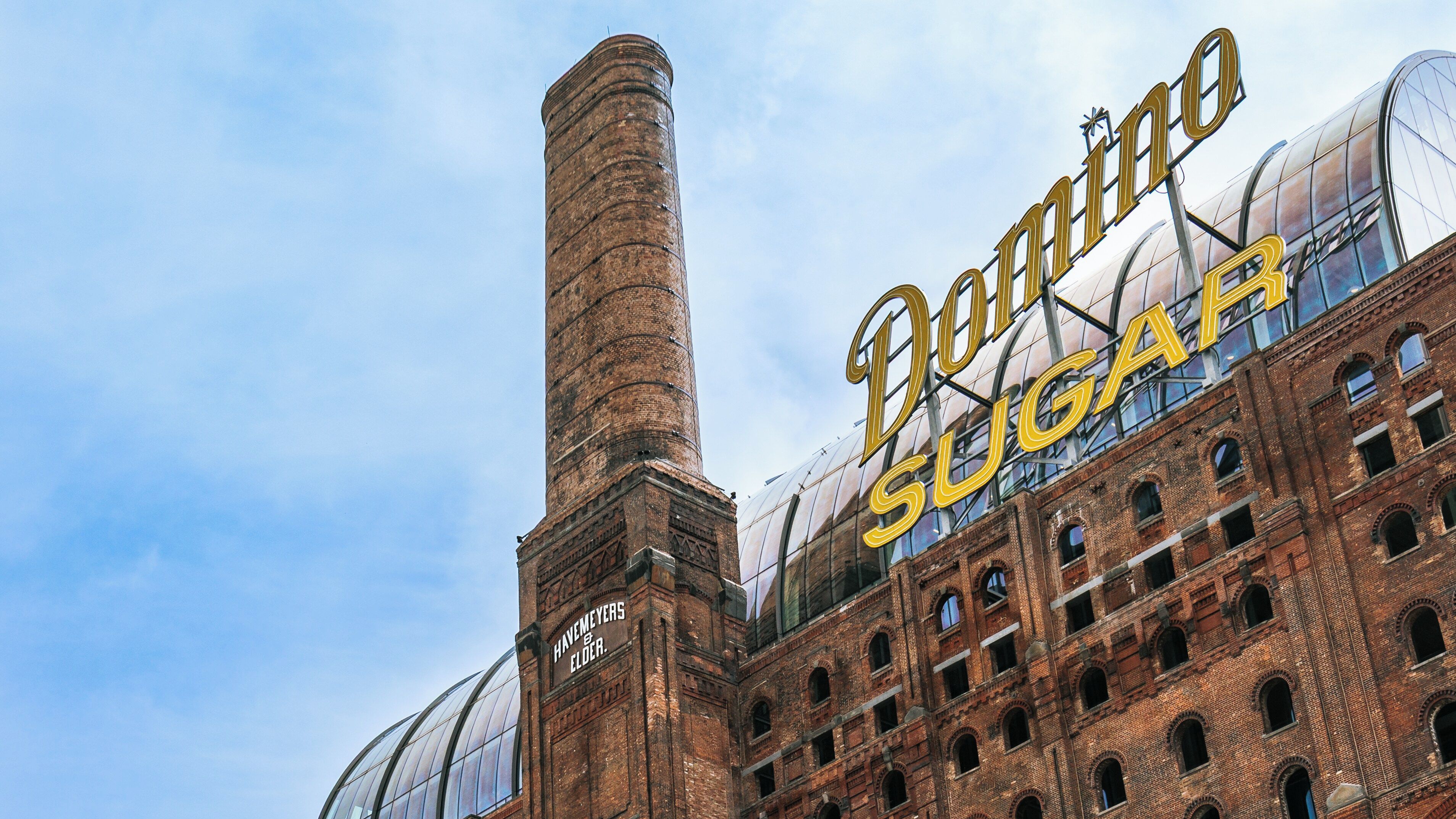 Ein historisches Backsteingebäude mit einem hohen Schornstein und einem großen Schild mit der Aufschrift 'Domino Sugar'.