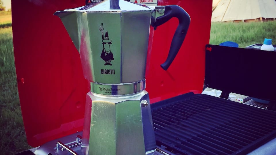 A portable stove with a pot on it, a tent in the background, and a grill.