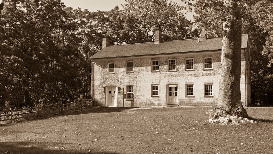 A large brick building with multiple windows and a central door, surrounded by trees and a grassy area.