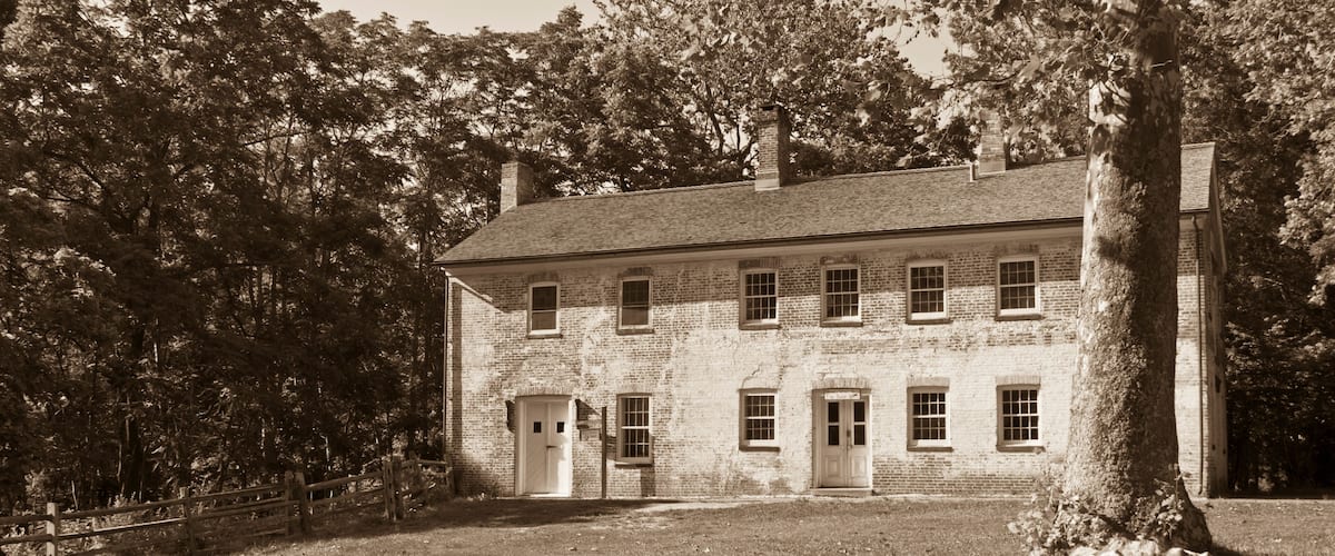 A large brick building with multiple windows and a central door, surrounded by trees and a grassy area.