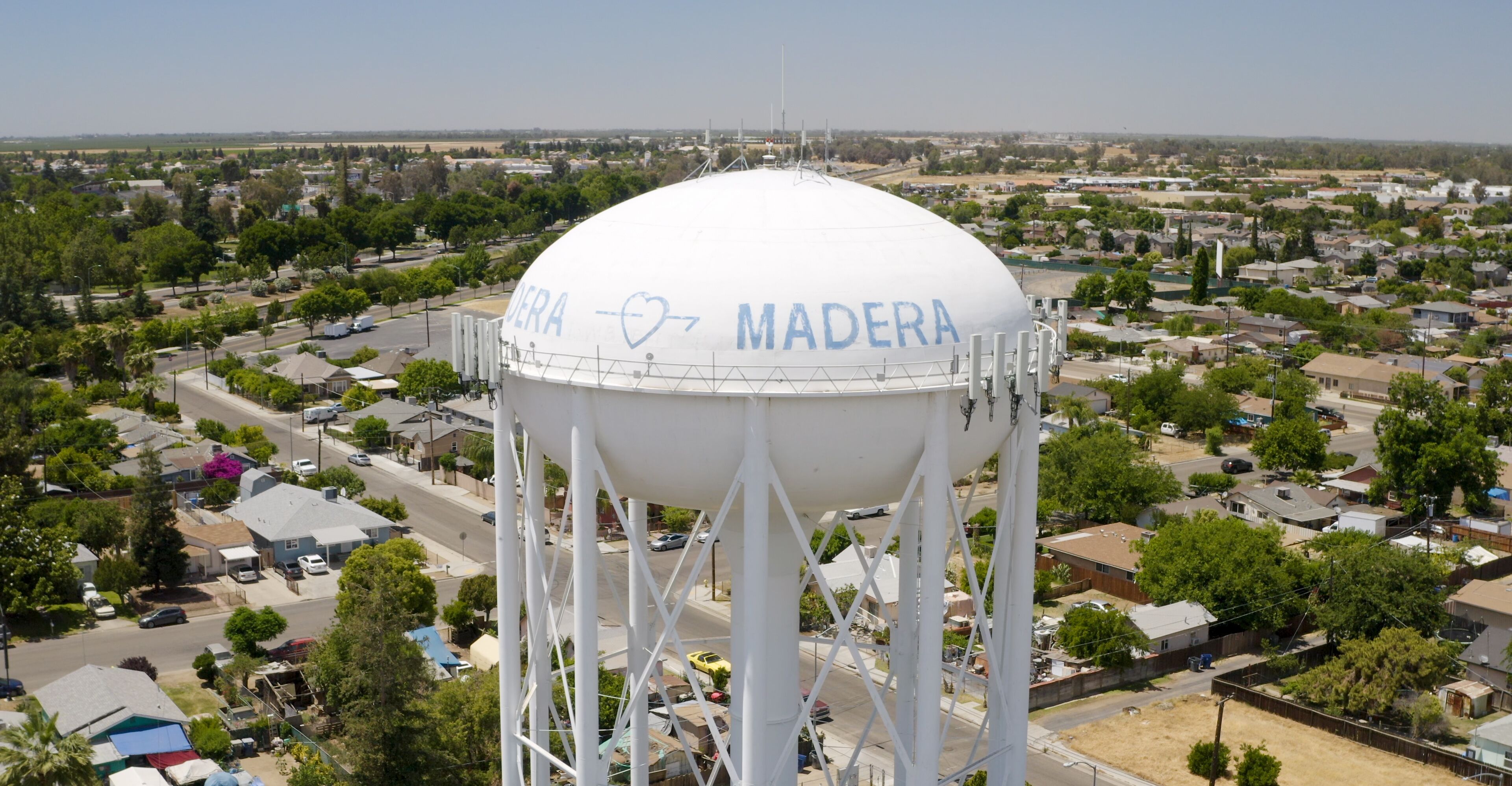 A water tower with 'DEERA MADERA' written on it, surrounded by residential houses and greenery.