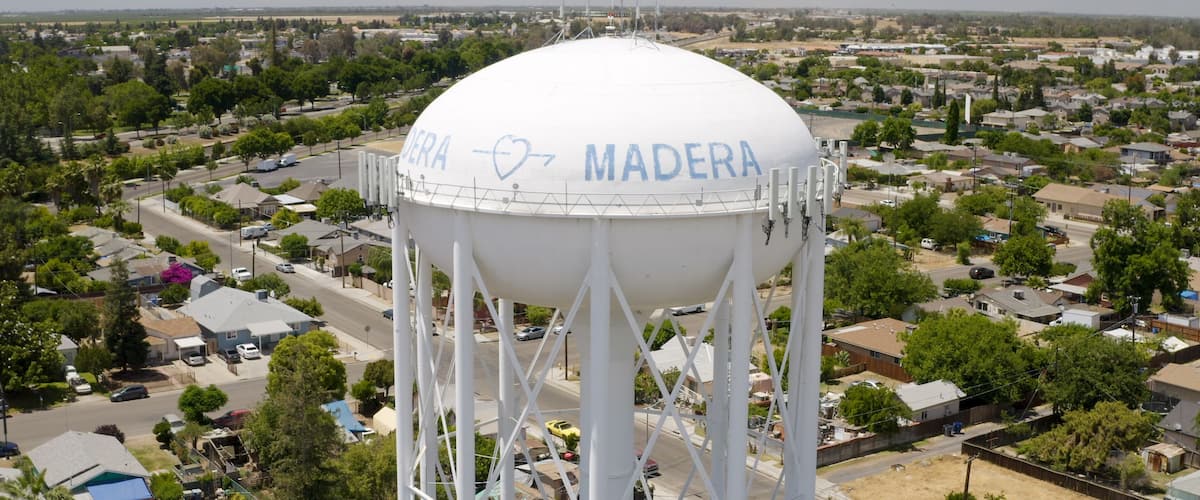 A water tower with 'DEERA MADERA' written on it, surrounded by residential houses and greenery.