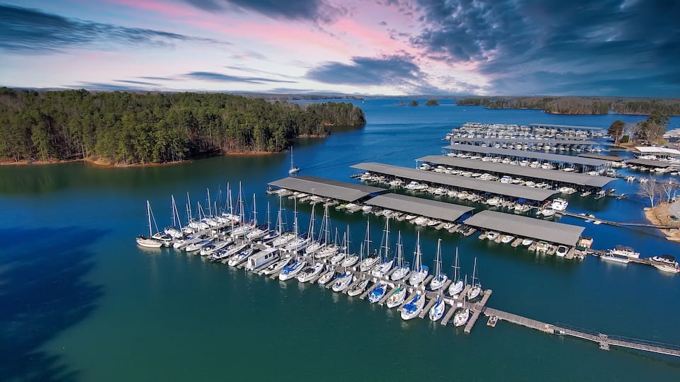 An aerial view of a marina with numerous boats docked, surrounded by a forested shoreline and a clear sky.