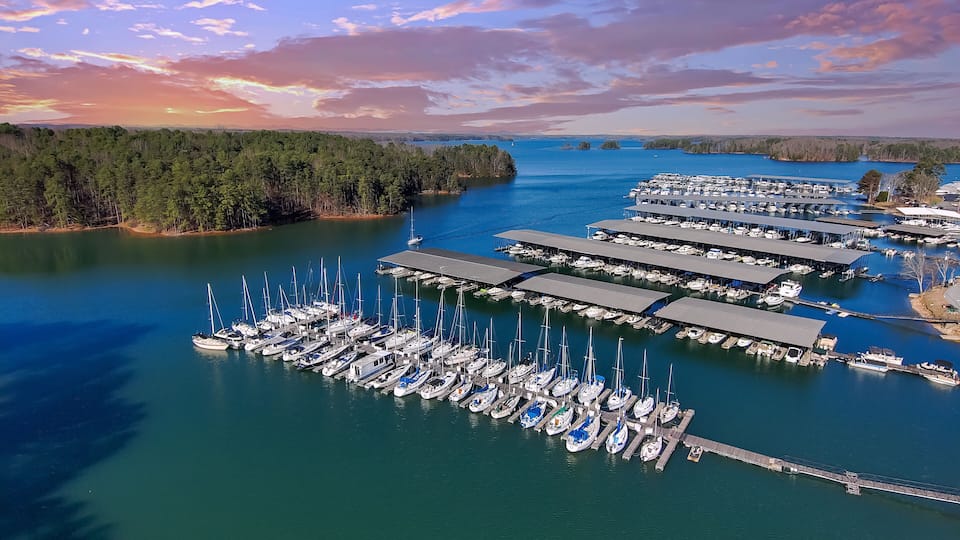 An aerial view of a marina with numerous boats docked, surrounded by a forested shoreline and a clear sky.