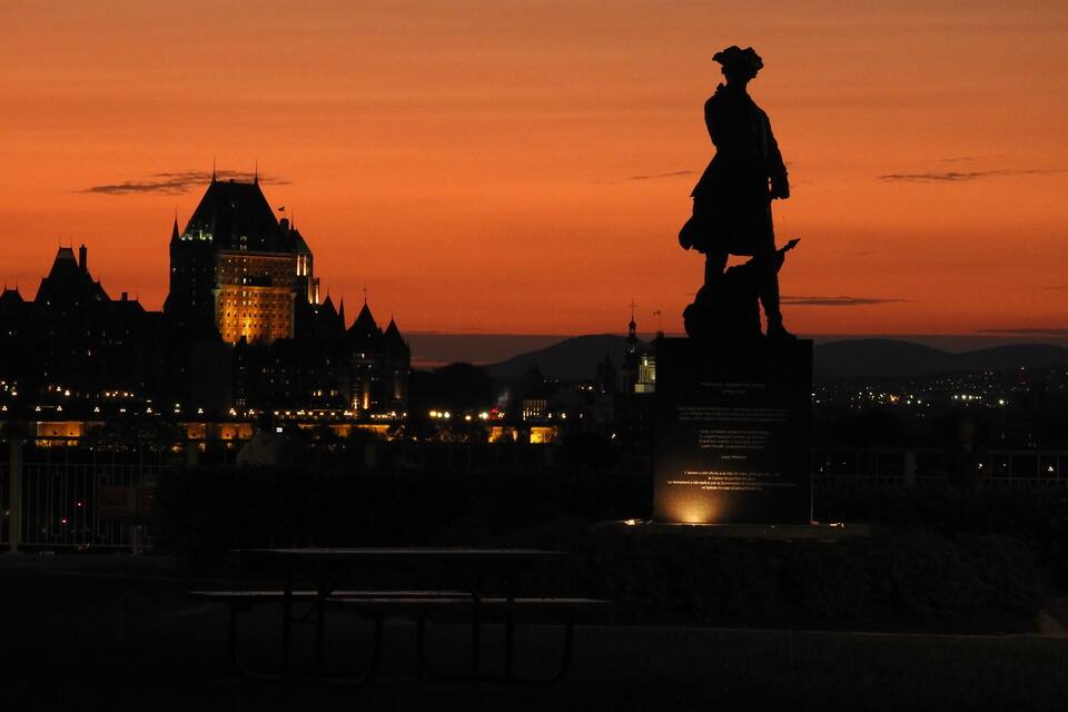 La silhouette d’une statue se détachant sur un ciel au coucher du soleil, avec un bâtiment historique en arrière-plan.