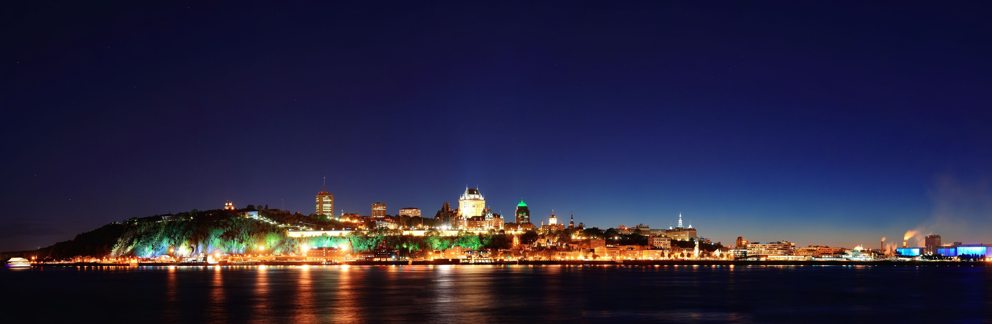 A cityscape at night with illuminated buildings and a river in the foreground.