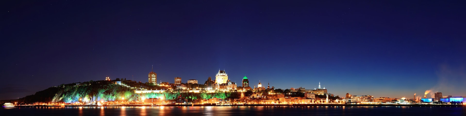 A cityscape at night with illuminated buildings and a river in the foreground.