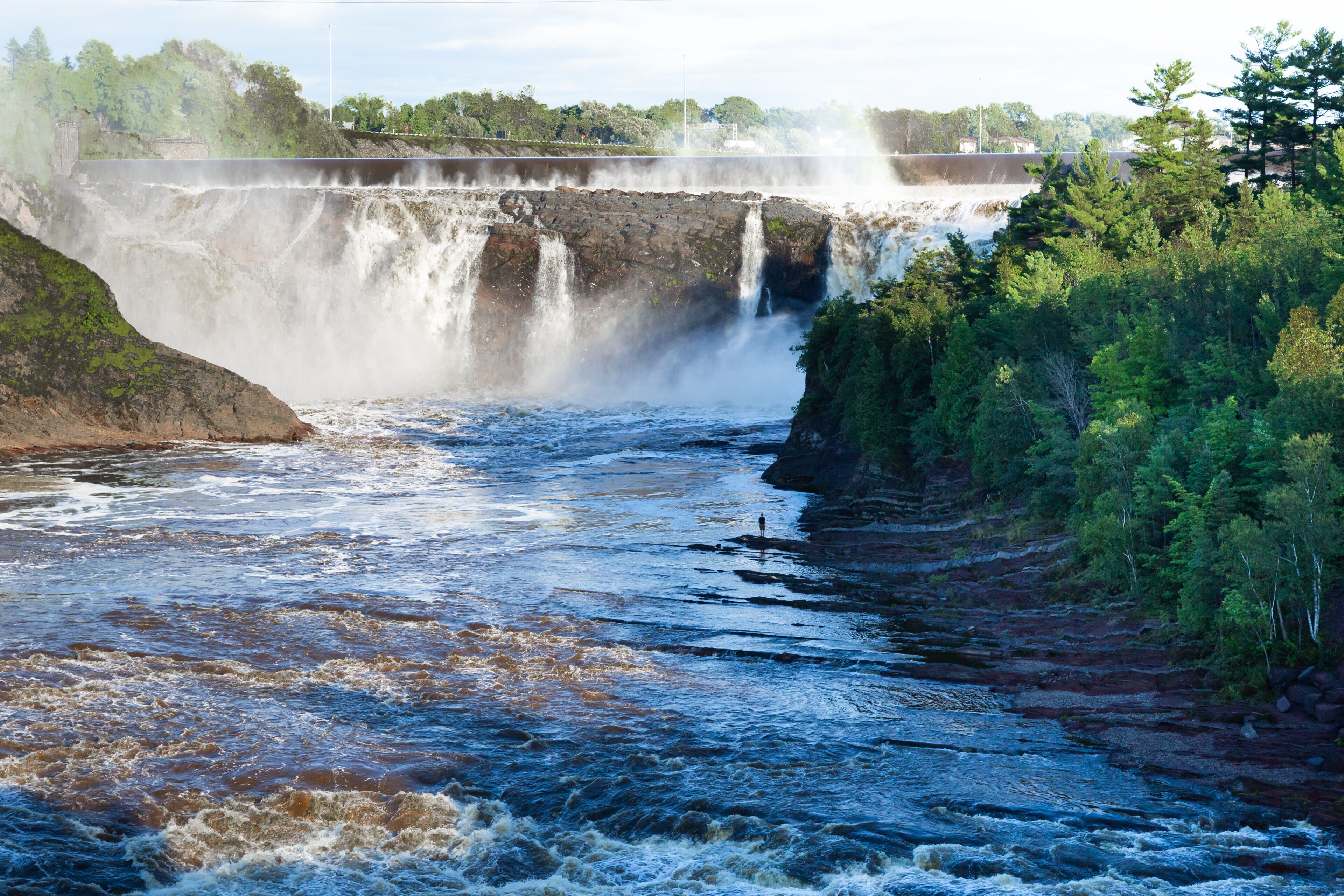 Tiny silhouette of young man standing on the shore of the Chaudière River during a summer late afternoon, with the famous falls in the background, Lévis, Quebec, Canada