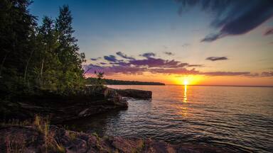 A sunset over a calm lake with rocky shores and trees.