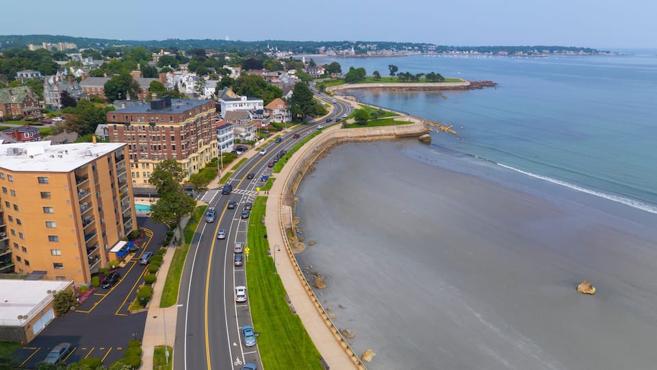 A coastal cityscape with a curved road along the beach, residential buildings, and a distant shoreline.