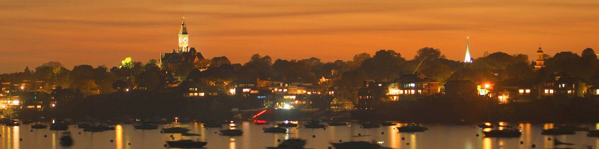 A city skyline at dusk with illuminated buildings, a prominent tower, and a calm harbor with boats.
