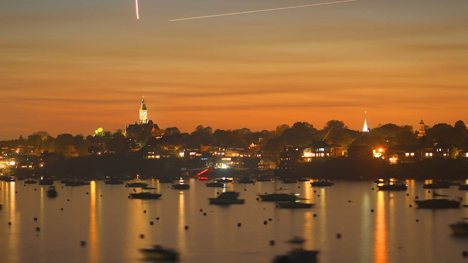 A city skyline at dusk with illuminated buildings, a prominent tower, and a calm harbor with boats.