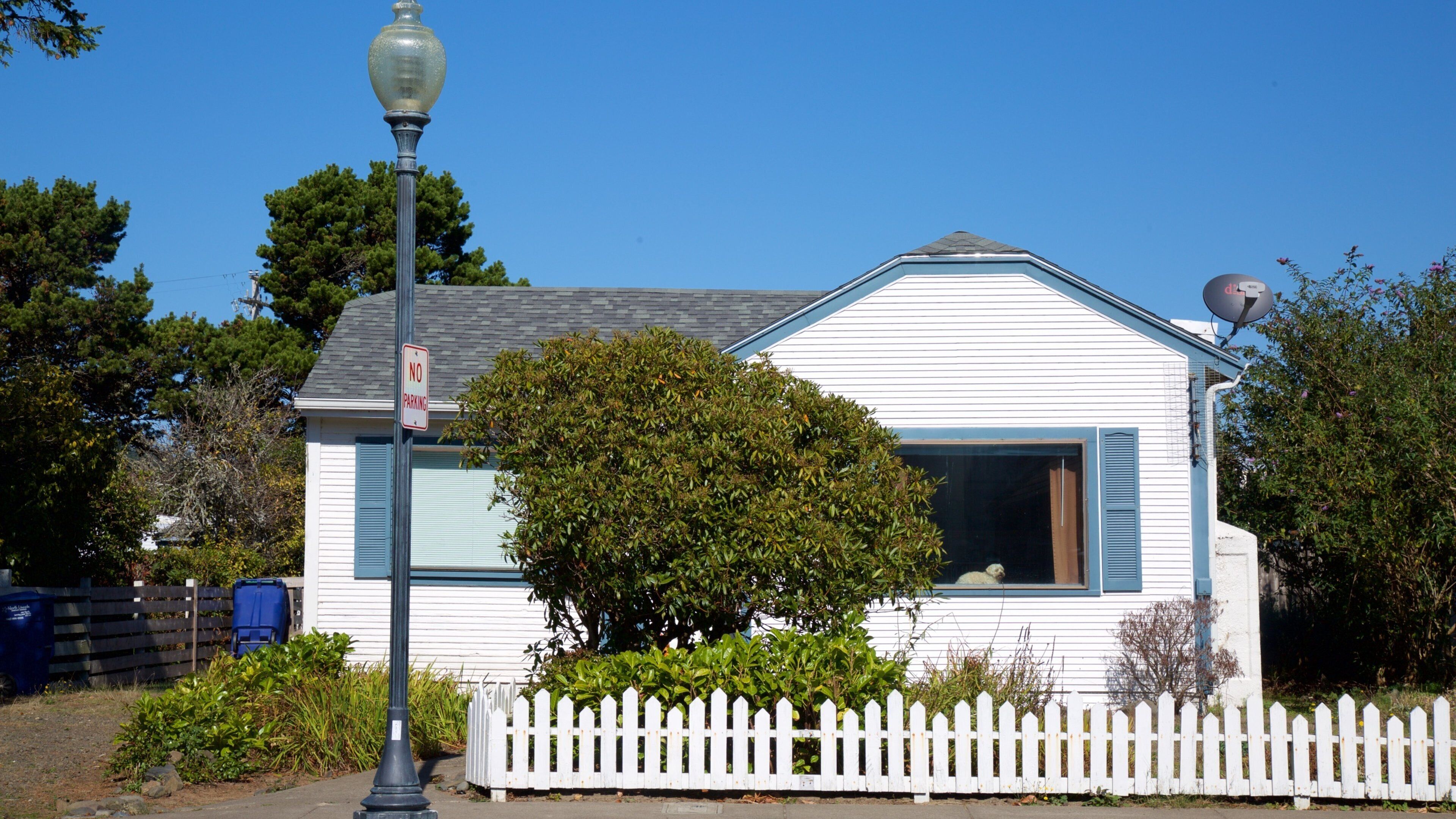 A white house with blue shutters, a white picket fence, and a 'No Parking' sign.