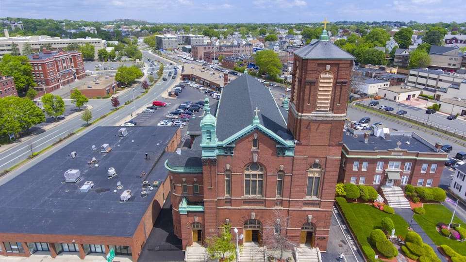 A large brick church with a green roof and a bell tower, surrounded by a parking lot and a busy street.