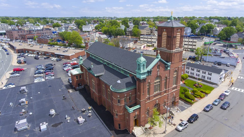 A large brick building with a green roof and a tower, surrounded by a parking lot and a cityscape.