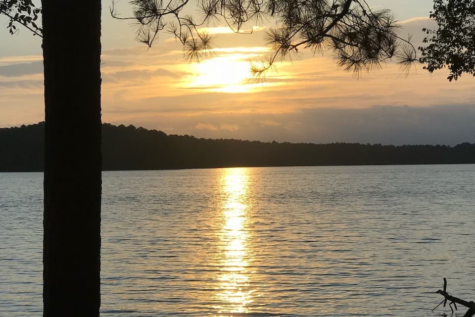 A sunset over a calm lake with reflections of the sun on the water, surrounded by trees and a distant shoreline.