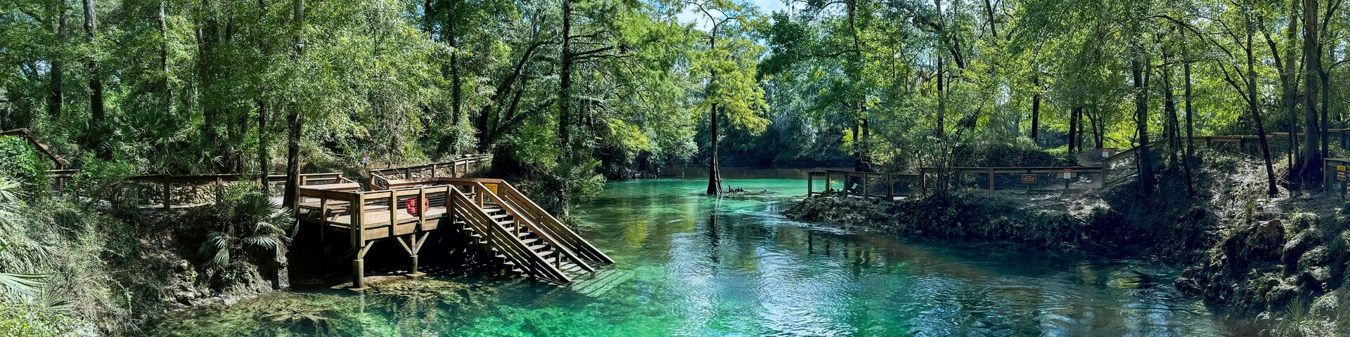 A clear river with wooden walkways and a bridge, surrounded by dense forest.