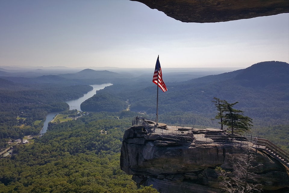 A river winding through a forested valley with a rocky outcrop and an American flag on top.