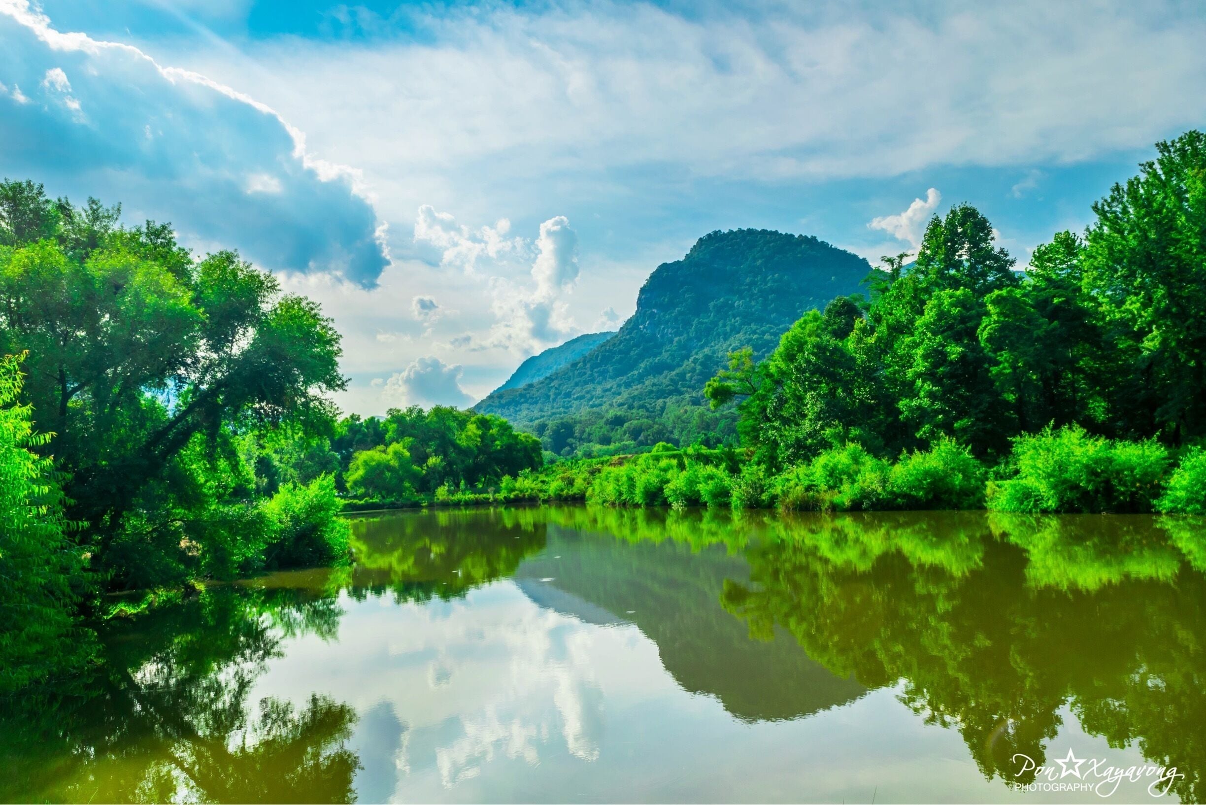 A calm river reflecting the surrounding greenery and a mountain in the background.