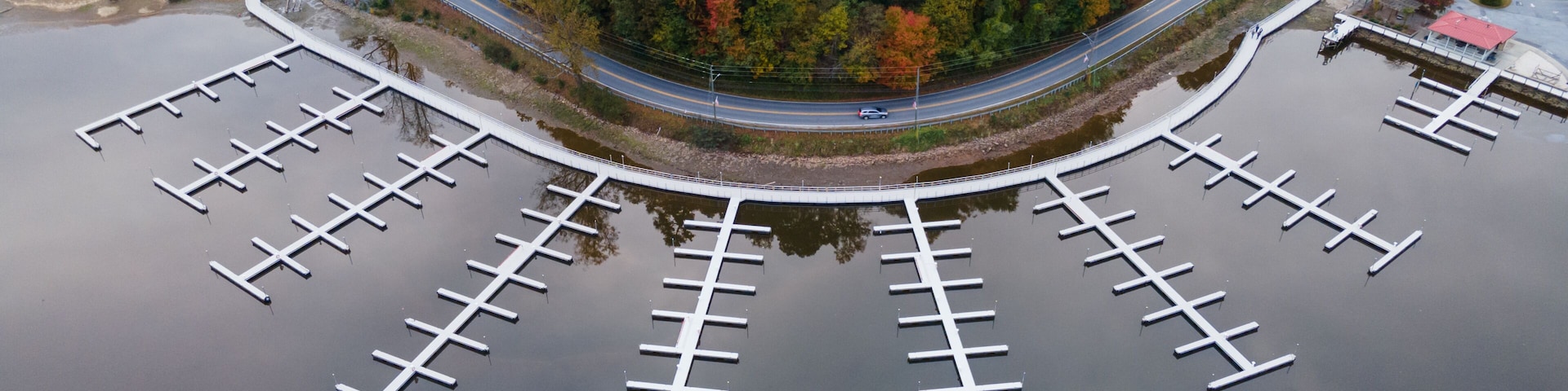 An aerial view of a marina with docks, surrounded by autumn-colored trees and a road curving around it.