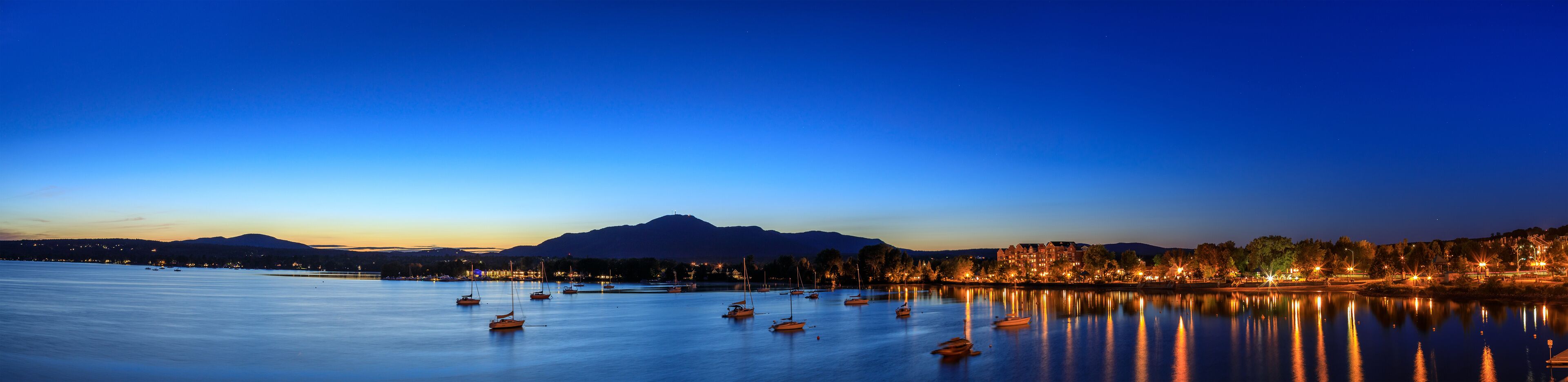 A calm lake at dusk with boats, a mountain in the background, and a lit-up town on the right.
