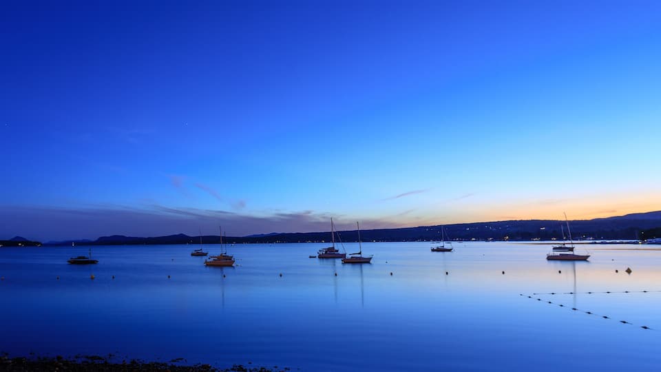 A calm harbor at dusk with several sailboats anchored in the water, a lighthouse on the left, and a mountain range in the background.