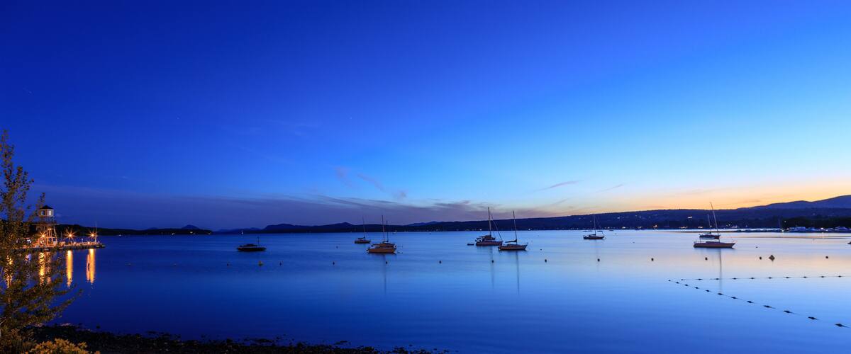 A calm harbor at dusk with several sailboats anchored in the water, a lighthouse on the left, and a mountain range in the background.