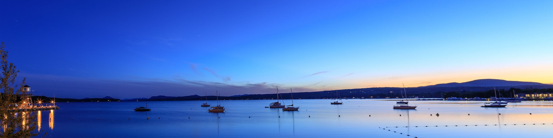 A calm harbor at dusk with several sailboats anchored in the water, a lighthouse on the left, and a mountain range in the background.