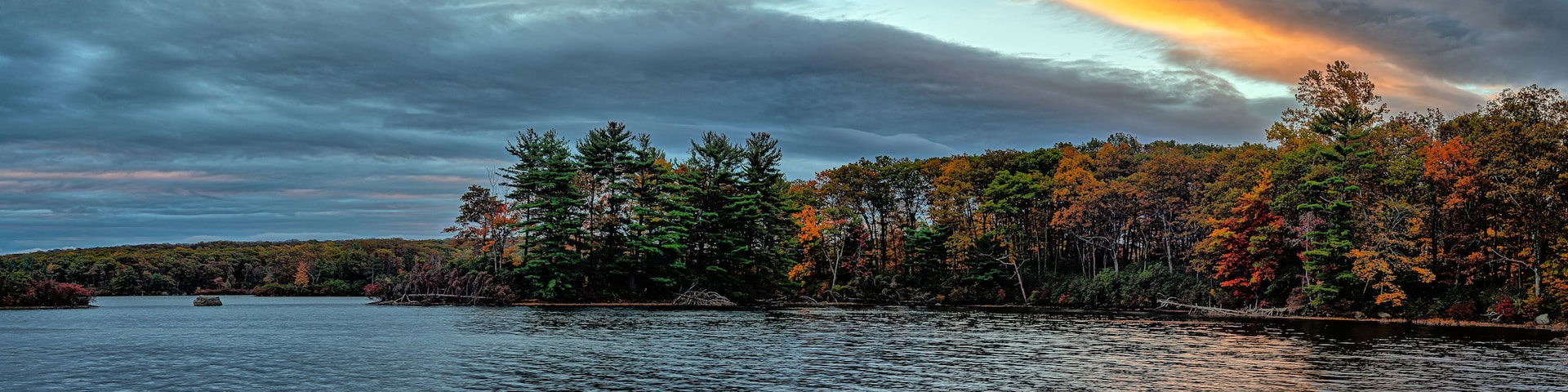 Un lac entouré d’arbres aux couleurs automnales, sous un ciel nuageux.