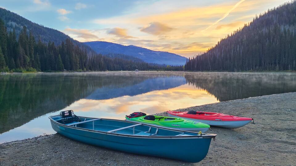 A calm lake with kayaks on the shore, surrounded by dense forest and mountains.