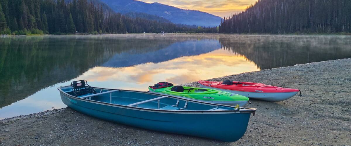 A calm lake with kayaks on the shore, surrounded by dense forest and mountains.