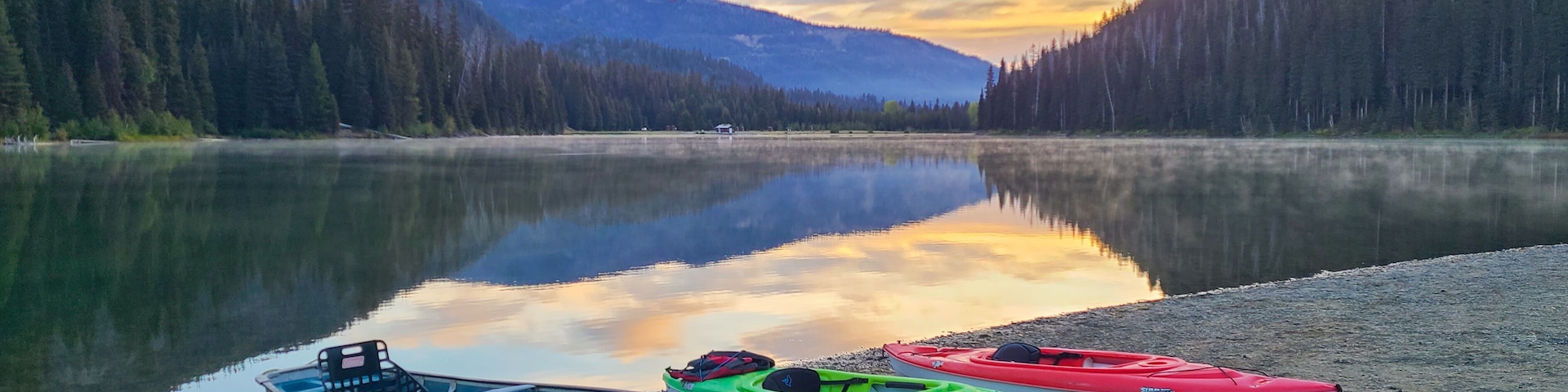 A calm lake with kayaks on the shore, surrounded by dense forest and mountains.
