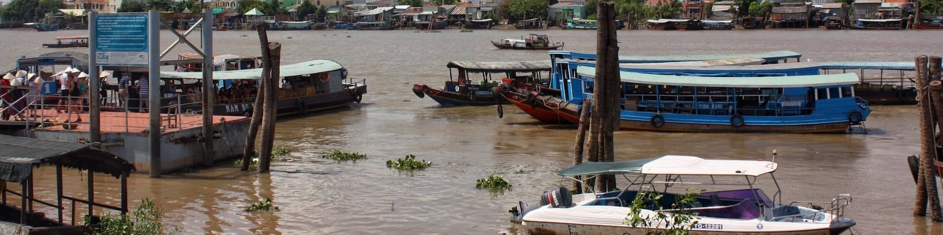 A river with boats docked along the shore, surrounded by buildings and palm trees.