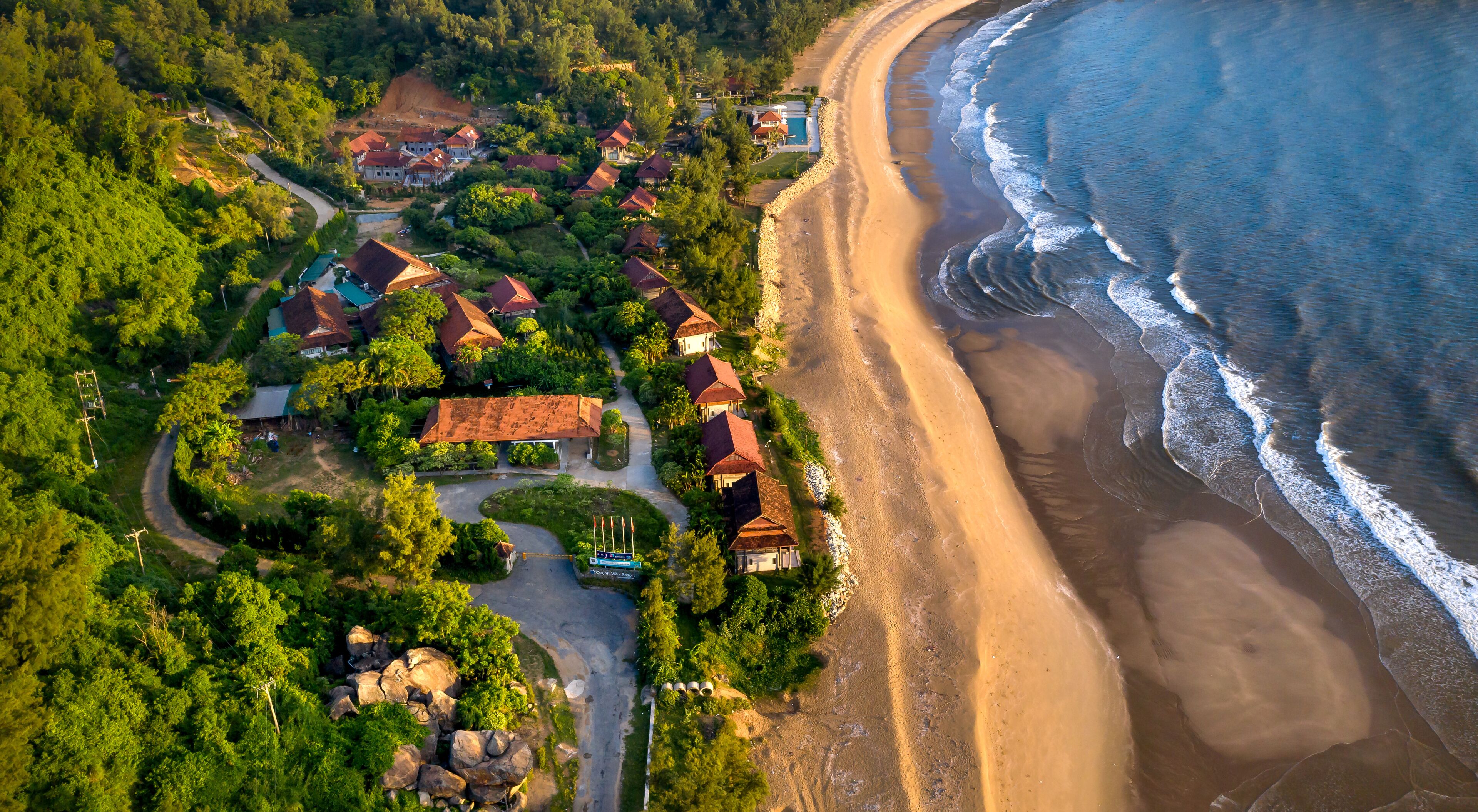 An aerial view of a coastal village with houses, a beach, and dense greenery.