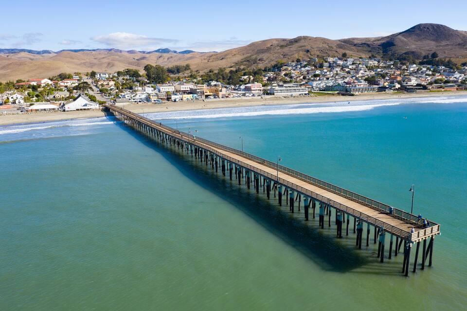 A long pier extending into a clear blue ocean, with a town and hills in the background.
