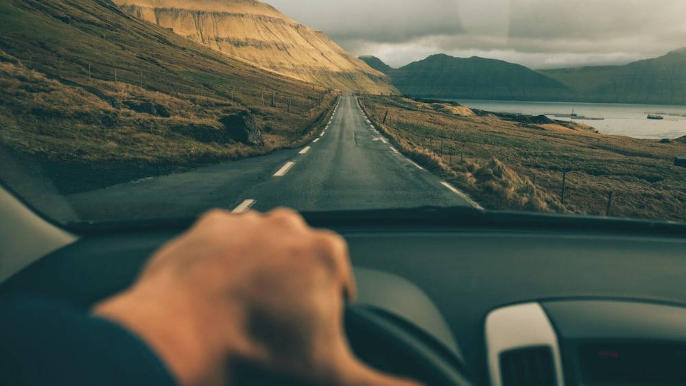 Eine Straße führt durch eine hügelige Landschaft, im Hintergrund ist ein Gewässer zu sehen.