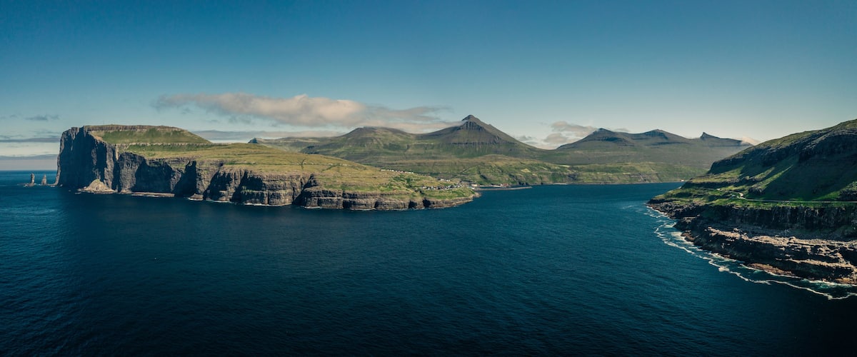 Panoramic view on coastline of island Eysturoy with rock formations Risin og Kellingin on Faroe Islands during the day with sun and blue sky, blue ocean and green cliffs.