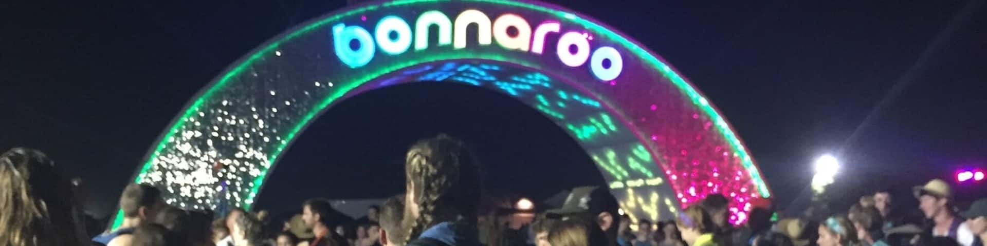 A crowd of people at an outdoor event with a large illuminated archway displaying the word "bonnaroo".