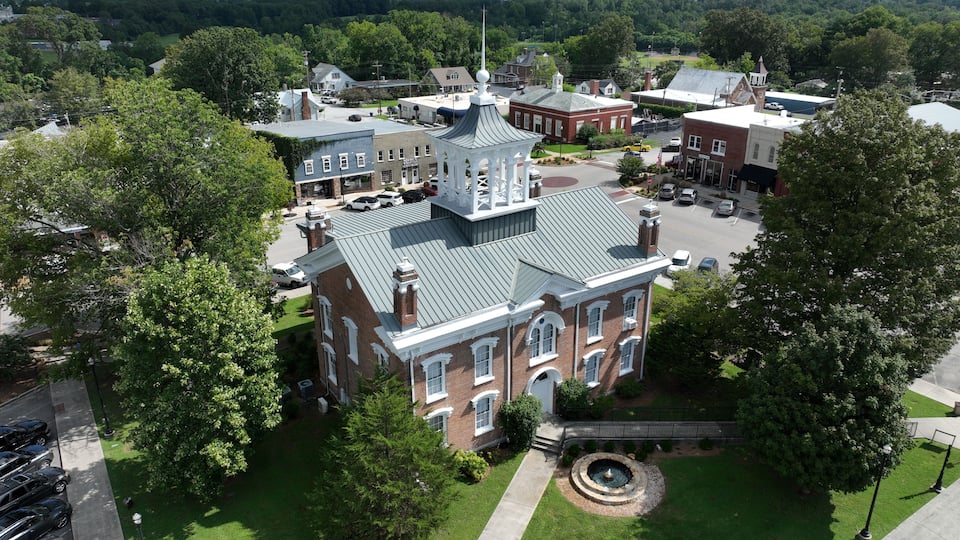 A large brick building with a clock tower, surrounded by trees and a small circular fountain.