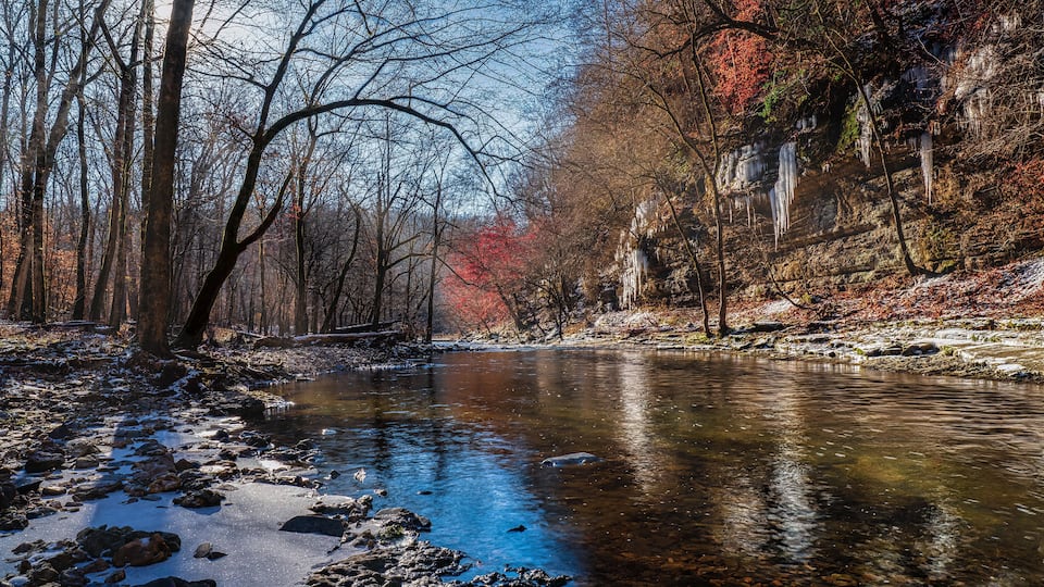 A river with clear water, surrounded by trees with autumn foliage, and icicles hanging from a cliff.