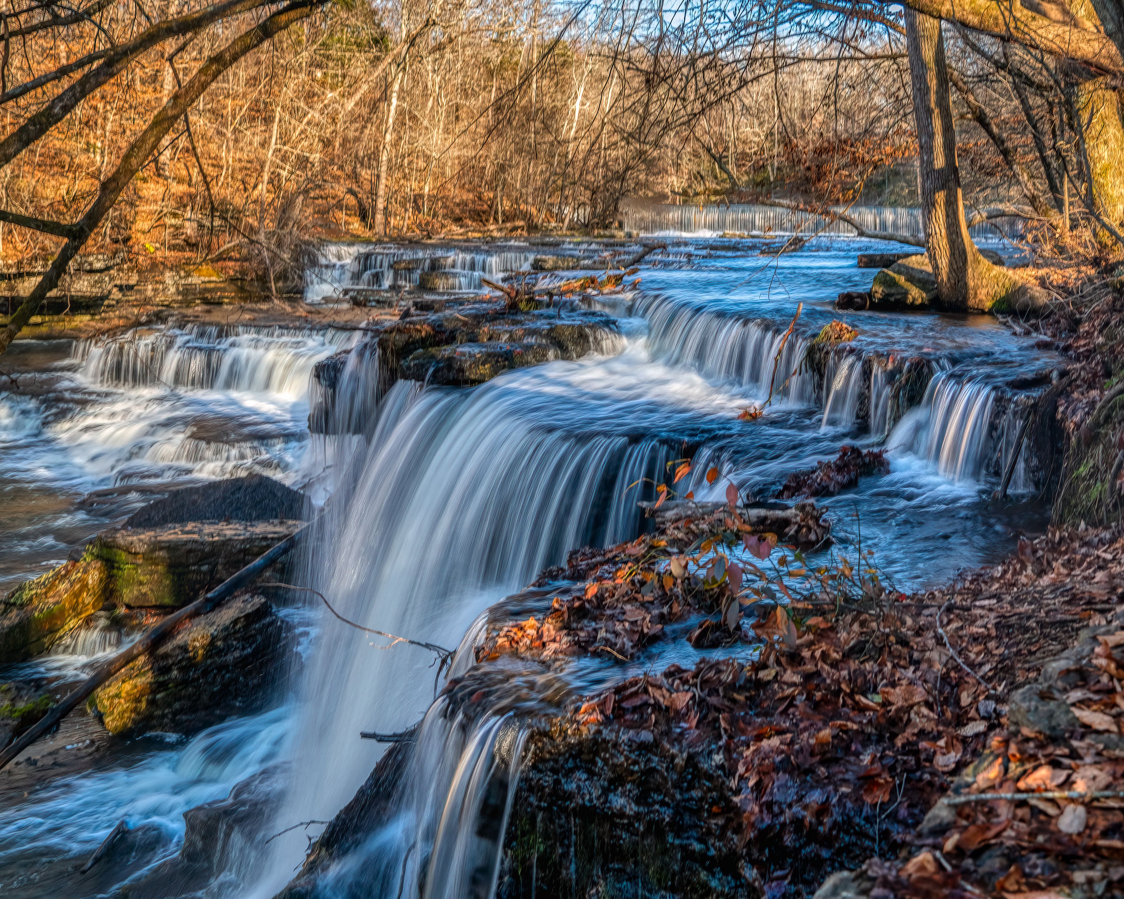 A multi-tiered waterfall in a forested area with autumn leaves.