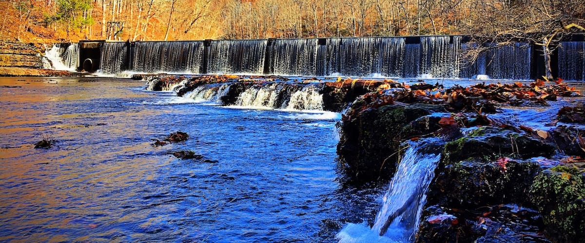 A waterfall cascading over rocks in a river, surrounded by autumn-colored trees and a clear blue sky.