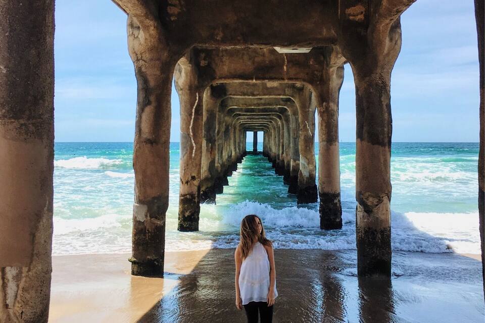 A person standing under a pier with a view of the ocean.