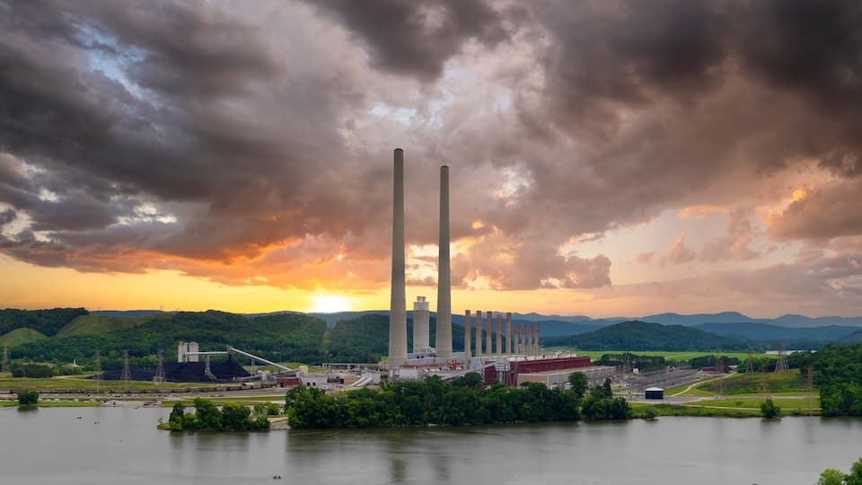 A power plant with multiple smokestacks, surrounded by a lake and green hills.