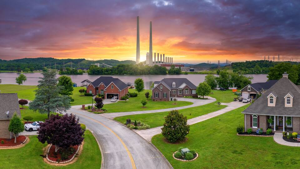 A suburban neighborhood with houses, a winding road, and a large industrial plant with tall chimneys in the background.